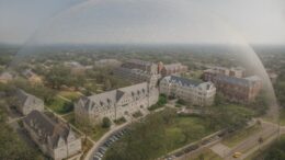 A hazy aerial view of Tulane University’s campus enclosed beneath a giant translucent dome, with the dome’s geometric pattern visible across the sky while campus buildings, lawns, and surrounding streets sit clearly underneath it.
