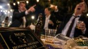 Close-up of a chaotic outdoor awards banquet table with an Neutral Ground News (NGN) Honor Awards plaque, spilled drinks, beads, and blurred guests celebrating in the background at night.