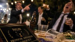 Close-up of a chaotic outdoor awards banquet table with an Neutral Ground News (NGN) Honor Awards plaque, spilled drinks, beads, and blurred guests celebrating in the background at night.