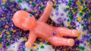 Close-up of a pink plastic king cake baby resting on white icing covered in purple, green, and gold sugar sprinkles.