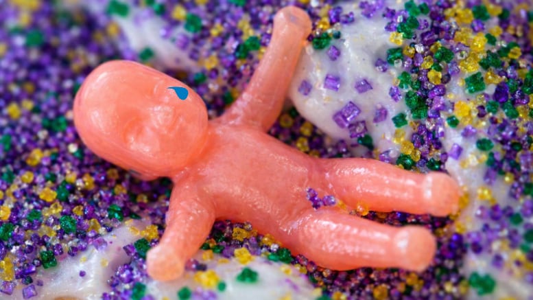 Close-up of a pink plastic king cake baby resting on white icing covered in purple, green, and gold sugar sprinkles.