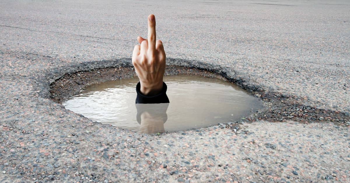 A pothole filled with water in the middle of a cracked street, with a human hand emerging from it, flipping the bird in frustration. New Study Finds 9 Out of 10 New Orleans Residents Have a Personal Vendetta Against a Specific Pothole