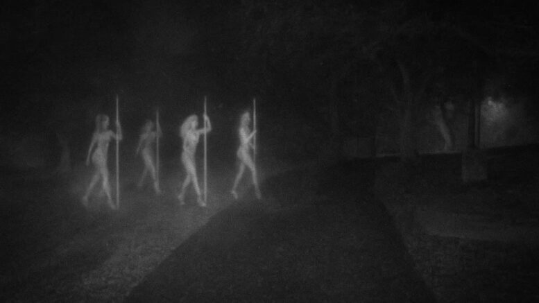 Grainy black-and-white image of a foggy nighttime park path with four ghostly, translucent female figures walking in a line while holding vertical poles. The eerie figures resemble spirits, with the background obscured by darkness and mist under looming oak trees.