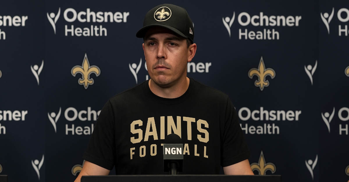 Kellen Moore speaks at a New Orleans Saints press conference, looking tired as he stands at the podium with an NGN microphone and Saints backdrop.