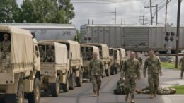 National Guard troops stand and sit beside a long convoy of military trucks stopped at a railroad crossing in Metairie, Louisiana, as a freight train blocks the road ahead. Some soldiers appear resting on the ground while others wait for the train to move.