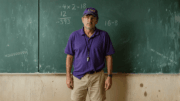 A middle-aged PE teacher wearing a wrinkled purple LSU shirt and a crushed LSU hat stands in front of a chalkboard with incorrect math and Gen Z insults like “MIDDDDDDDDD” and “bro thinks he’s him fr” written behind him.