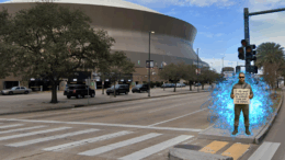 A man in futuristic clothing stands outside the Caesars Superdome in New Orleans holding a glowing sign that reads, “If you want civilization to survive, turn off the games,” surrounded by faint blue energy as cars pass by.