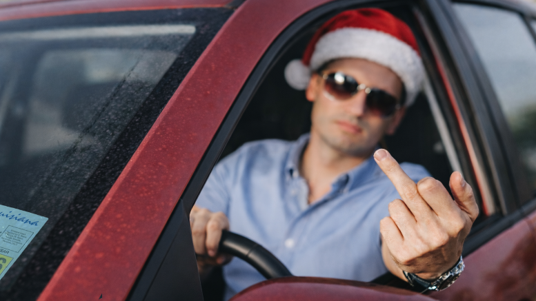 Man wearing a Santa hat and sunglasses gestures from inside a red car while seated behind the steering wheel.