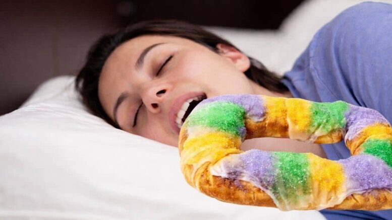 Woman sleeping in bed with a king cake positioned near her mouth, associated with Carnival season in Louisiana.