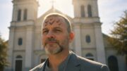 Bald man with multiple ash check marks on his forehead standing in front of St. Louis Cathedral in New Orleans on Ash Wednesday, sunlight behind him in an editorial-style portrait.