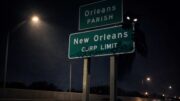 Nighttime view of New Orleans city limit and Orleans Parish highway signs on I-10, partially lit by a streetlight, with a dark shadowy figure lurking behind the sign.