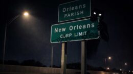 Nighttime view of New Orleans city limit and Orleans Parish highway signs on I-10, partially lit by a streetlight, with a dark shadowy figure lurking behind the sign.