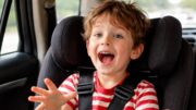 Animated 6-year-old child in a car seat talking excitedly during a car ride, gesturing mid-story in backseat.