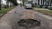 Handwritten sign in a New Orleans pothole reading “It isn’t NOLA without potholes support your local pothole” on a residential street.