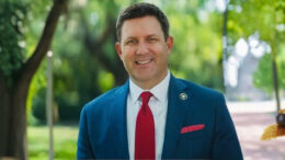 Jefferson Parish Councilman Scott Walker smiling in a suit with a small pot of gold in the background.