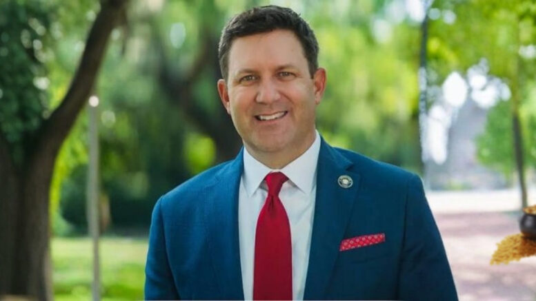 Jefferson Parish Councilman Scott Walker smiling in a suit with a small pot of gold in the background.