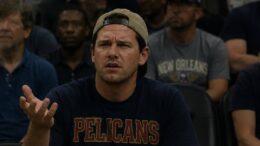 A frustrated New Orleans Pelicans fan sits in the arena crowd with one hand raised in confusion.
