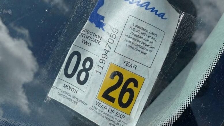 A partially peeled Louisiana brake tag curls away from a vehicle windshield as sunlight and clouds reflect across the glass.