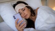 Woman wearing a sleep mask lying in bed while looking at her phone for a Permanent Daylight Saving Time article about Louisianans being "on my way."