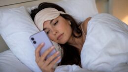 Woman wearing a sleep mask lying in bed while looking at her phone for a Permanent Daylight Saving Time article about Louisianans being "on my way."