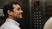 A man stands beside the elevator control panel with a subtle smile as he confidently assumes the unofficial role of pressing buttons for fellow passengers.