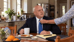 Mitch Landrieu sits at a kitchen table while his elderly mother adjusts his tie, accompanying a Neutral Ground News story about a possible presidential run.