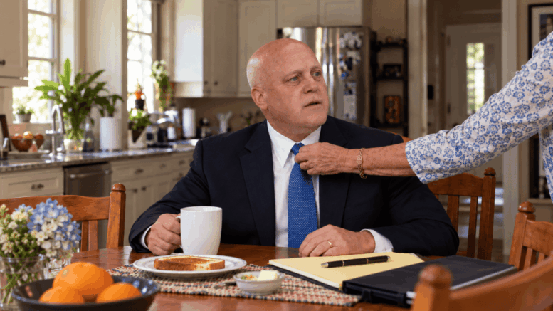 Mitch Landrieu sits at a kitchen table while his elderly mother adjusts his tie, accompanying a Neutral Ground News story about a possible presidential run.
